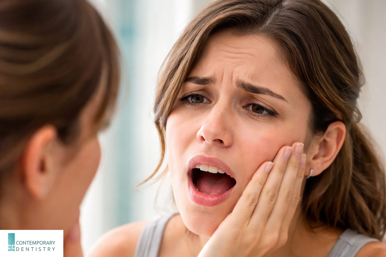 Woman touching her cheek while looking in a bathroom mirror with a painful expression, illustrating toothache or dental pain.
